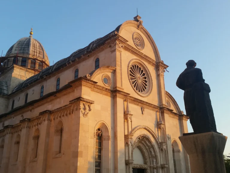 Cathedral of St James in Šibenik, its stone facade glowing in evening light beside a dark statue.