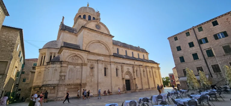 The stone Cathedral of St James fills a sunny square in Šibenik, with café tables and people in the foreground.