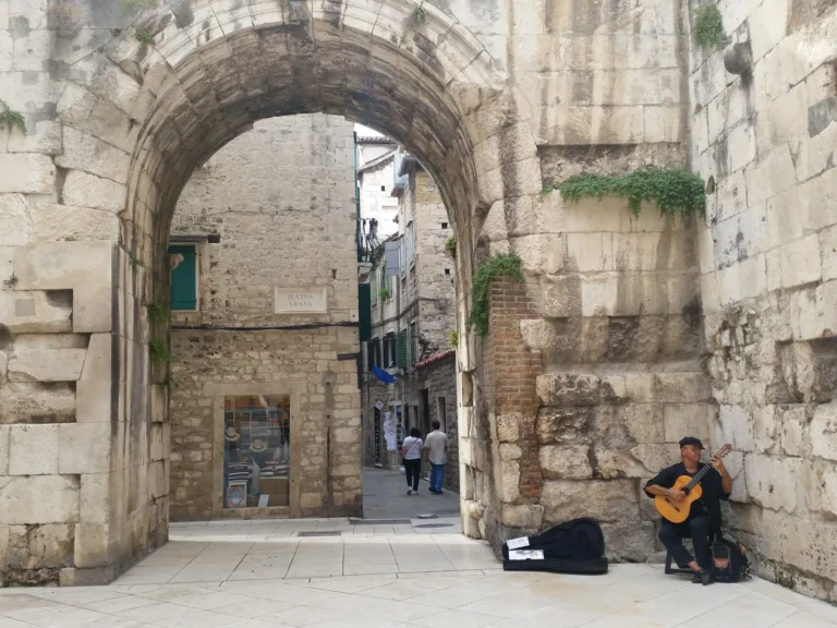 A street musician plays guitar beside Split’s stone Golden Gate, with the old-town lane visible through the arch.
