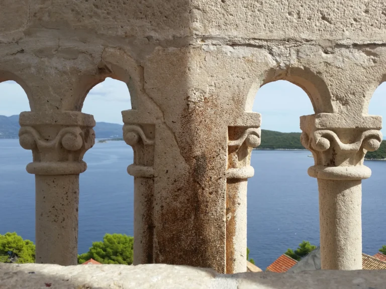 Carved stone arches frame a blue sea, distant green shoreline and red-tiled rooftops on the Croatian coast