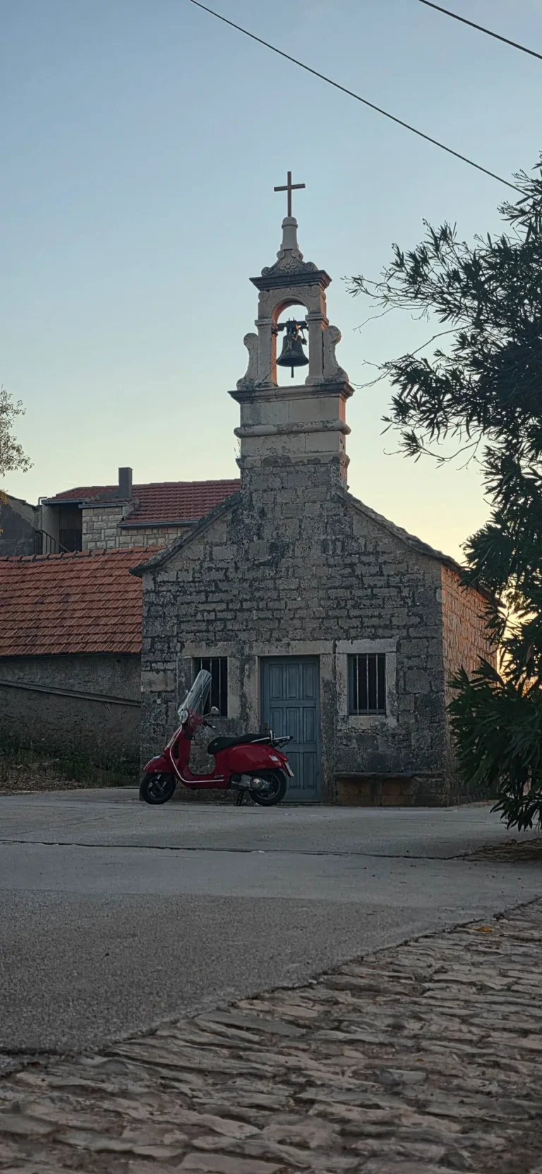 A small stone chapel with a bell tower and cross stands behind a parked red scooter at dusk.