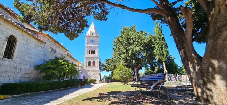 A stone church and clock tower beside a shaded garden with a bench.