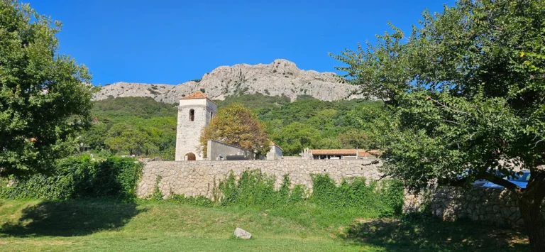 A stone church tower and low buildings beneath a rocky mountain ridge, framed by trees.