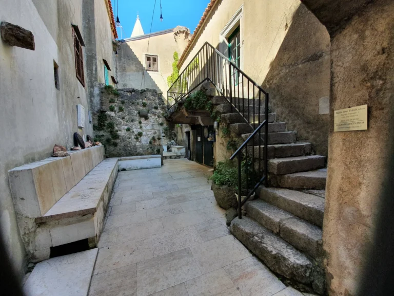 A narrow stone courtyard with a raised stone staircase, pale walls and a small tiled landing.