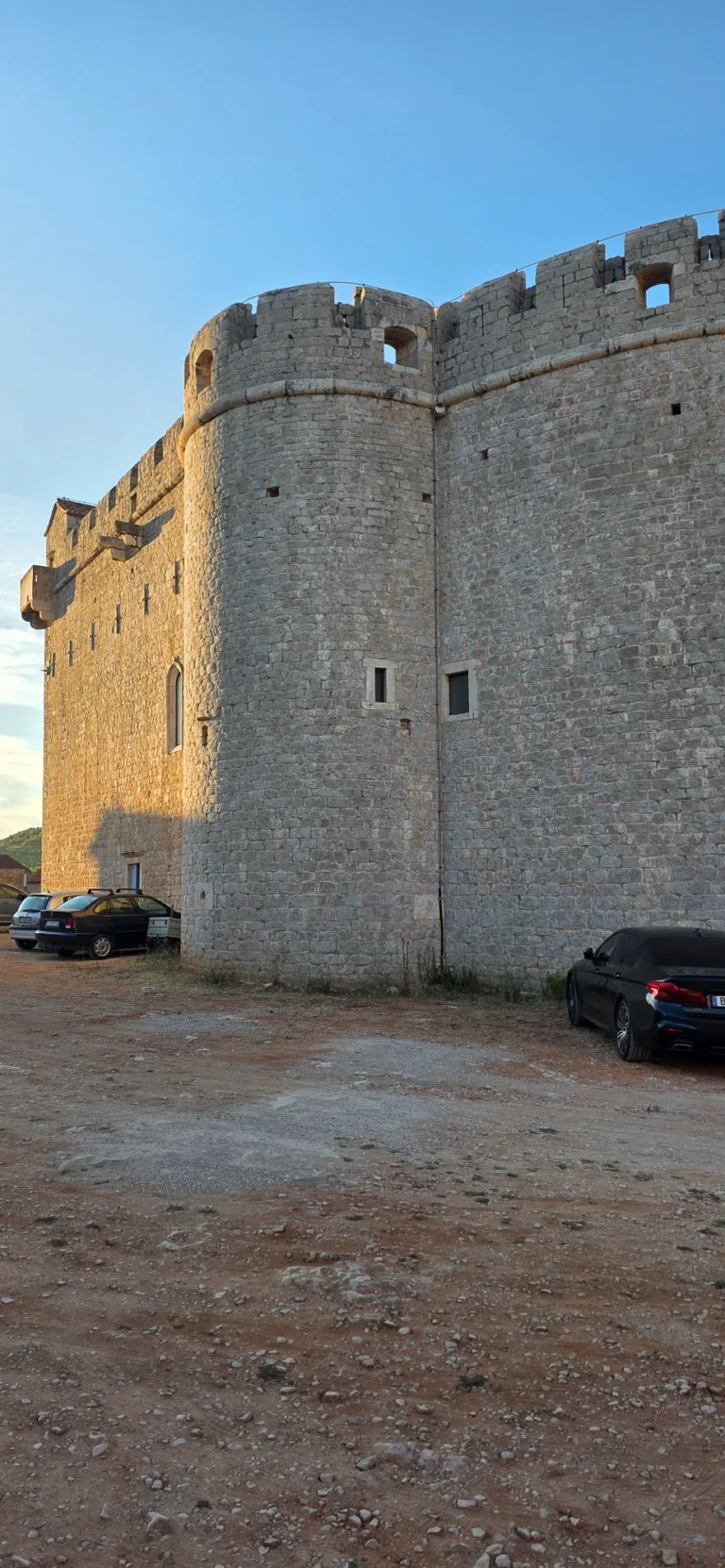 A massive stone fortress with a rounded tower stands beside a gravel parking area under a clear blue sky.