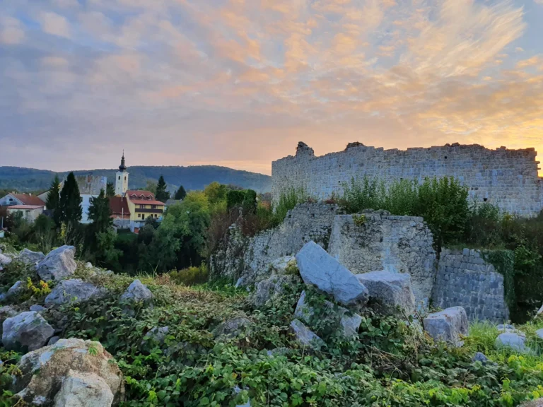 Stone fortress ruins above a village with a church tower beneath a glowing sunset sky