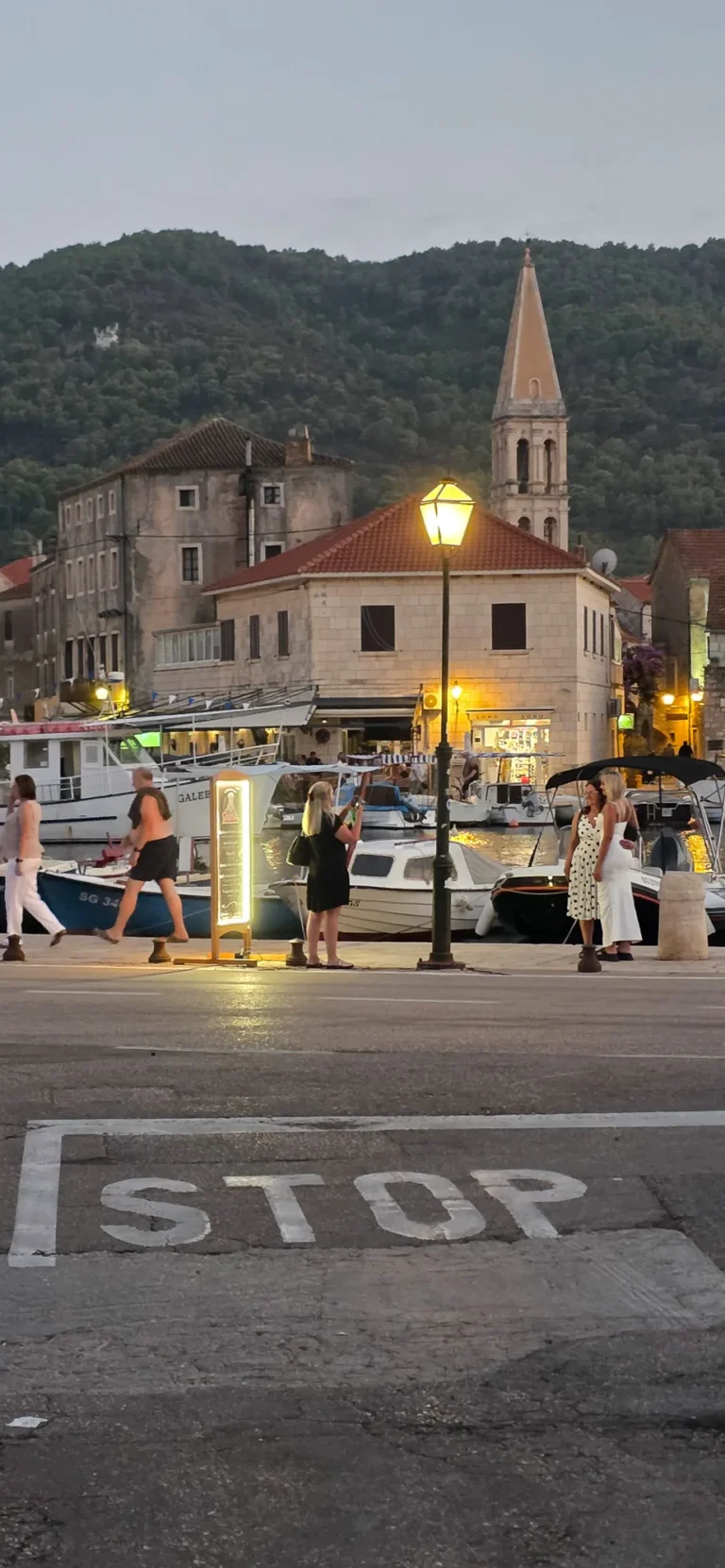 Boats and pedestrians at a stone harbour beneath a church bell tower at dusk.
