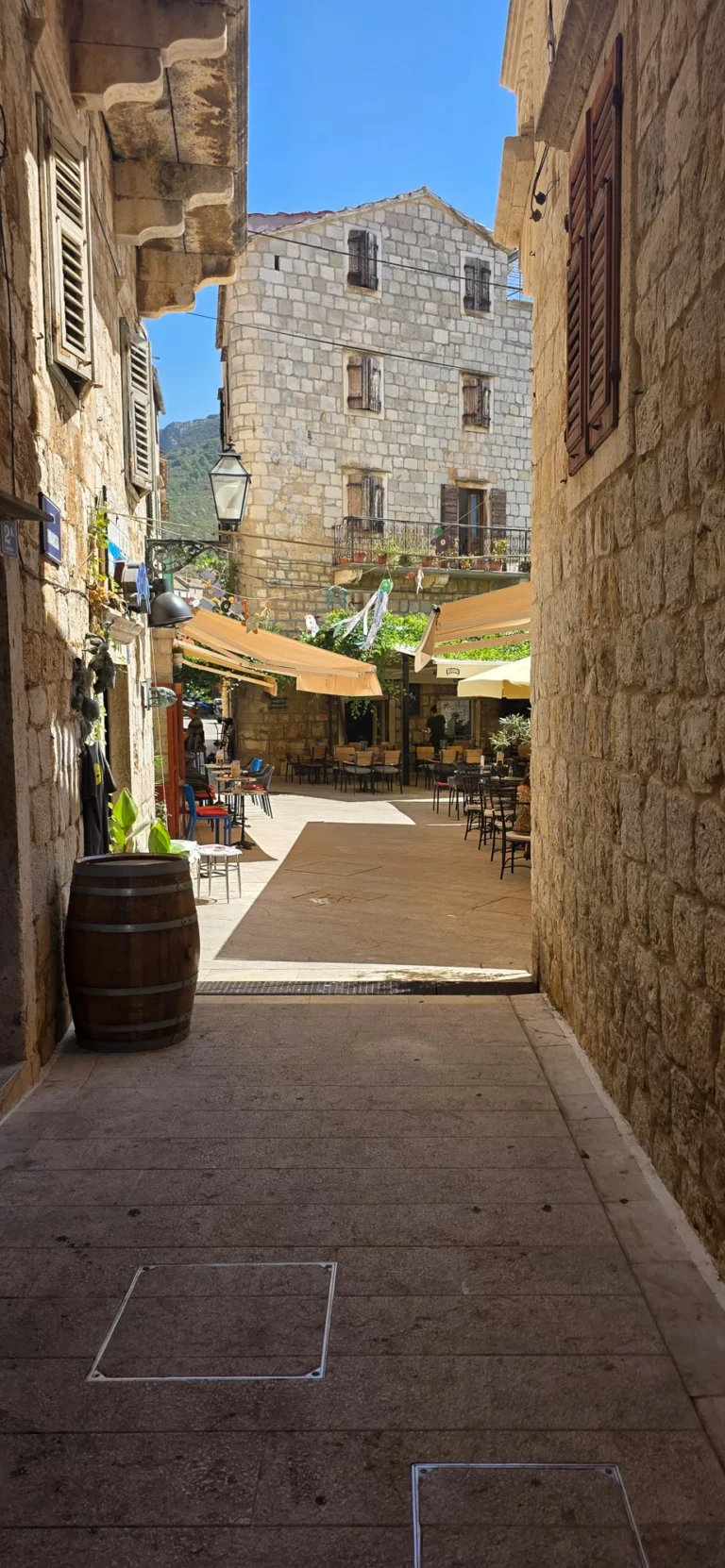 A narrow stone-paved alley opens onto a shaded café square with outdoor tables.