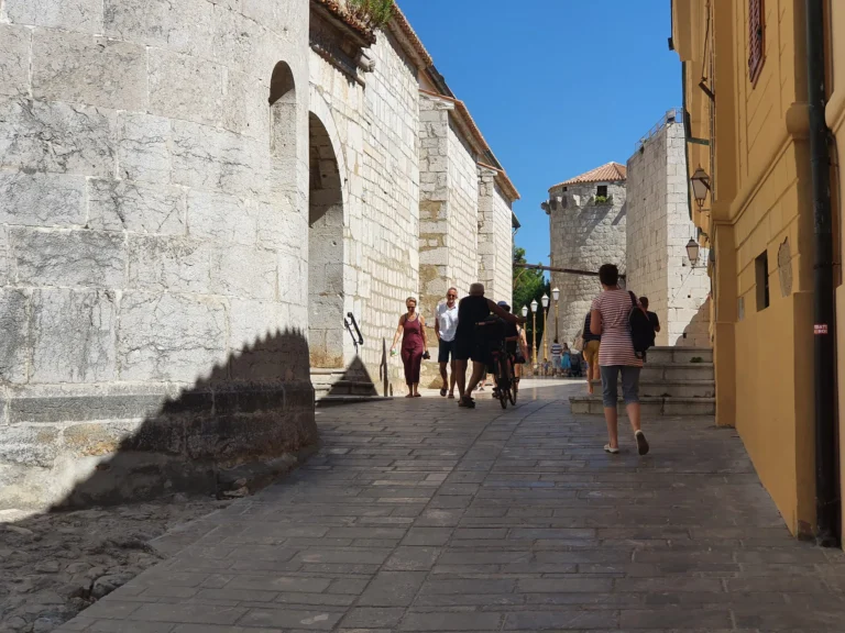 People walk along a paved stone street between pale stone buildings and an arched wall.