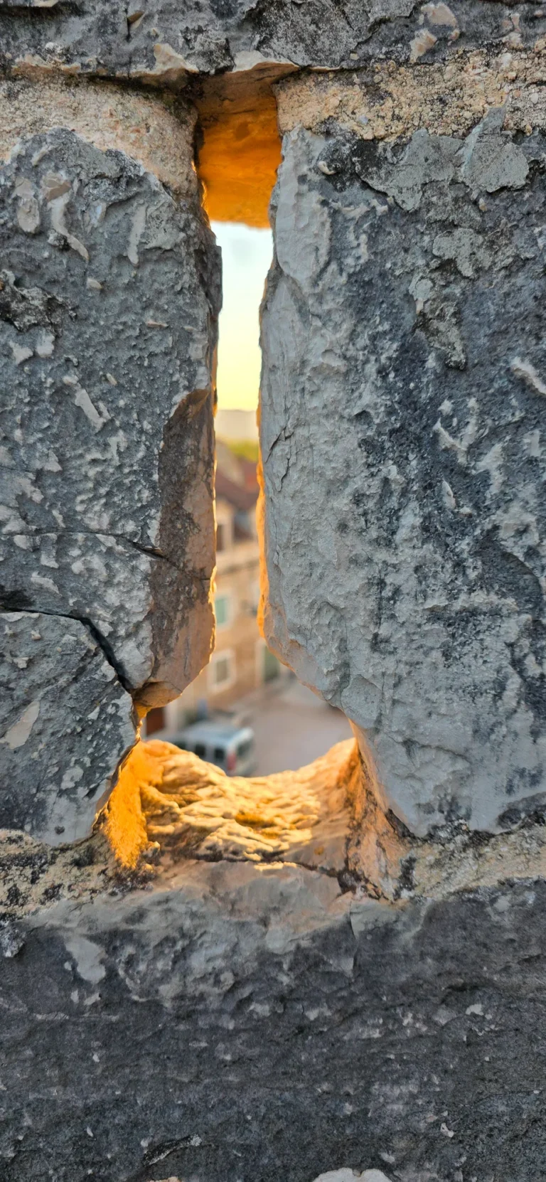 Warm evening light shines through a narrow opening in a rough stone wall onto a blurred street.