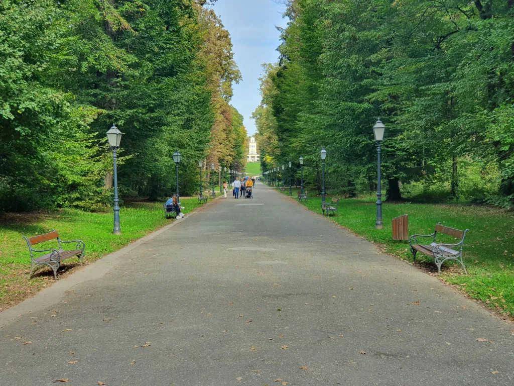 A broad park path runs between leafy trees, benches and traditional lampposts, with a few people walking in the distance.