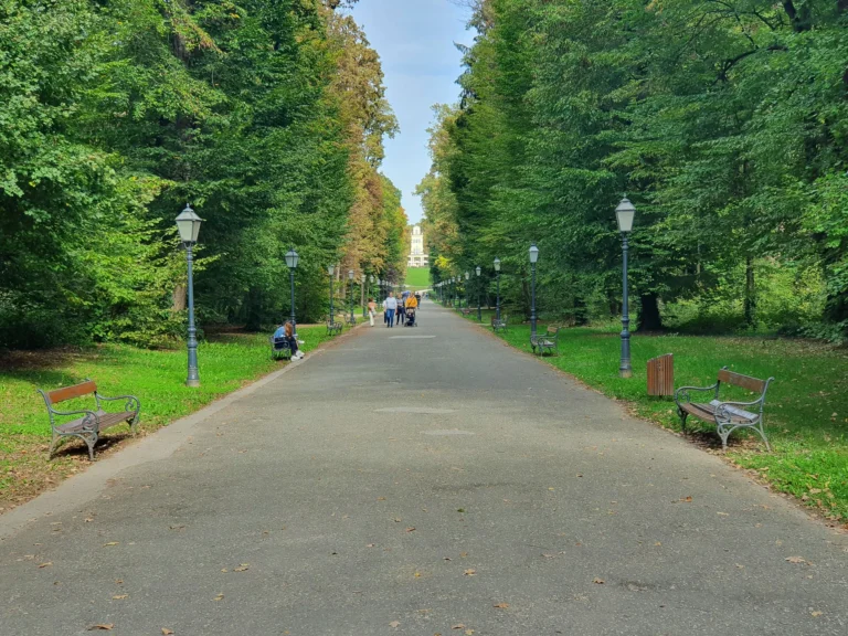 A broad park path runs between leafy trees, benches and traditional lampposts, with a few people walking in the distance.