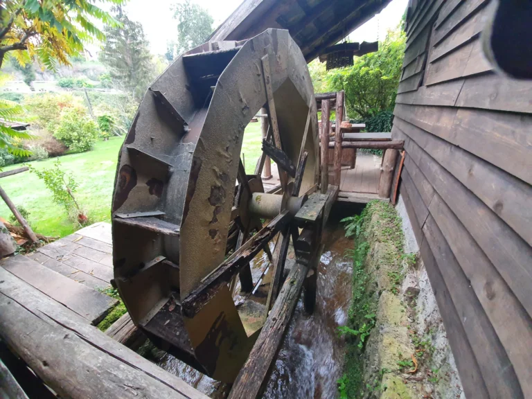 A large weathered waterwheel beside a wooden mill, with water flowing beneath it in a leafy garden.