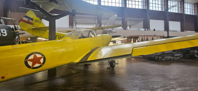 A yellow vintage aircraft stands beside other military vehicles in an indoor museum hall.