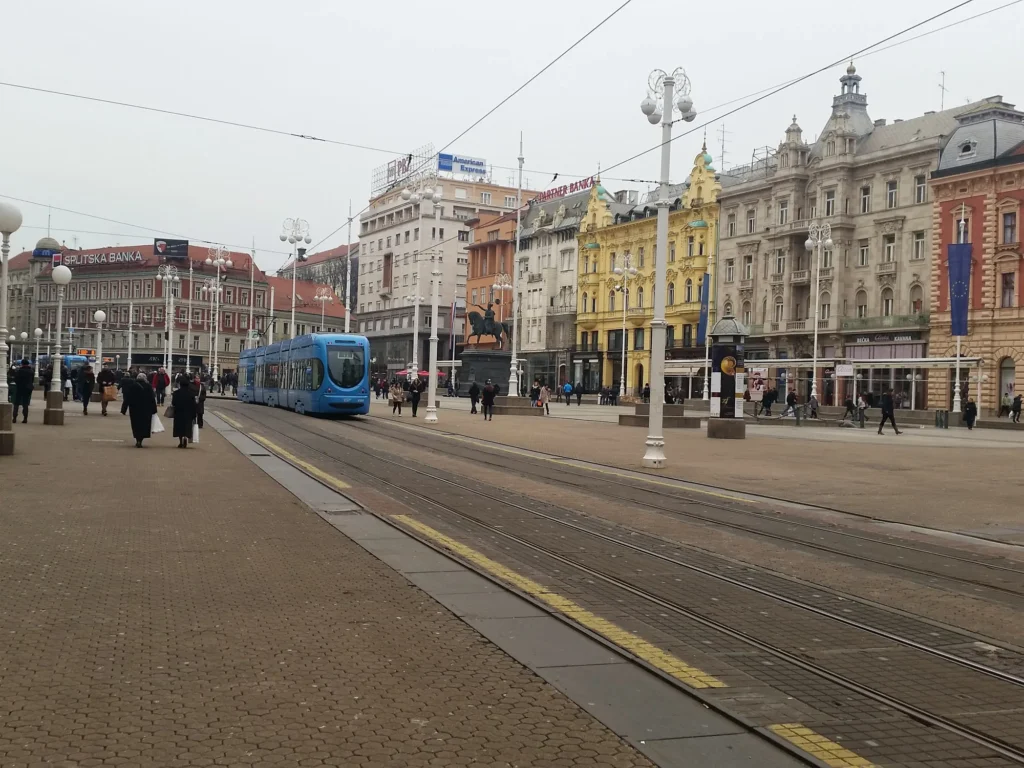 A blue tram crosses Ban Jelačić Square in Zagreb, surrounded by ornate city buildings and pedestrians.