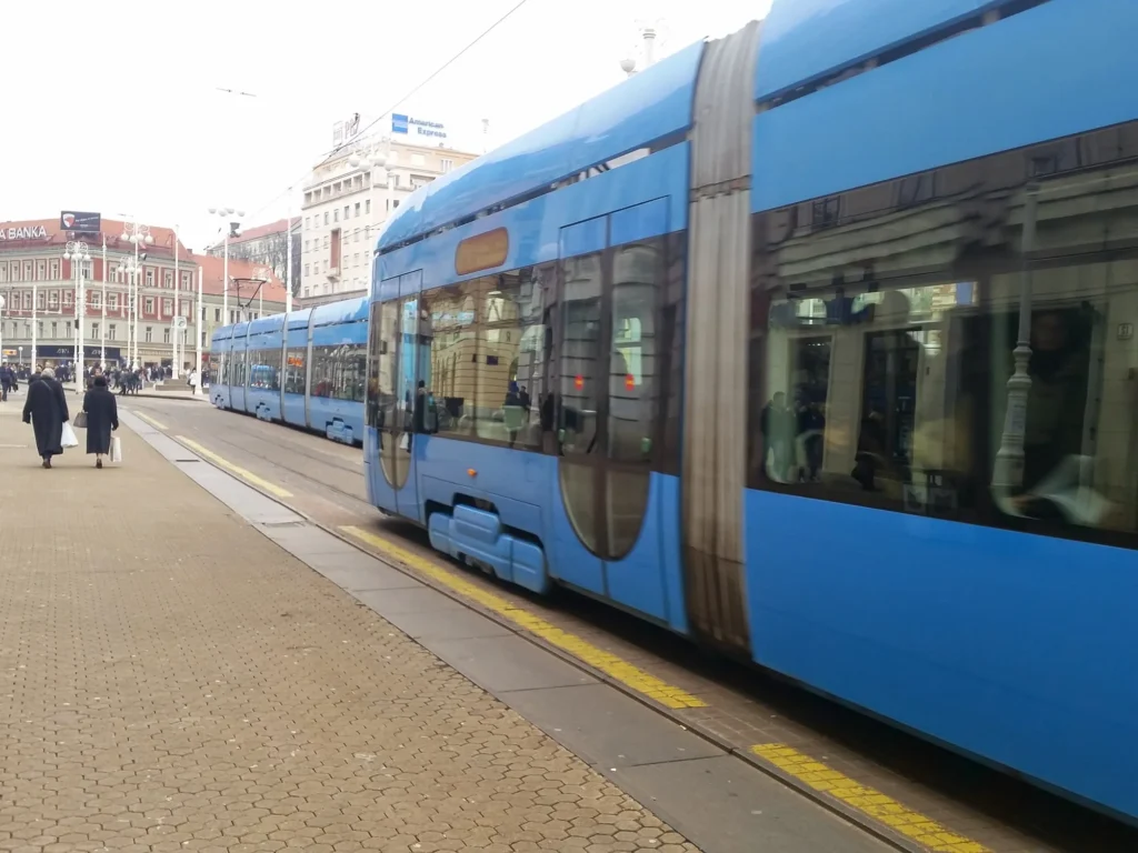 A blue tram passes pedestrians and historic buildings in Zagreb city centre.