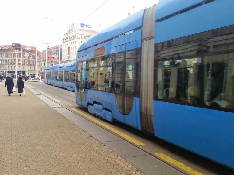 A blue tram passes pedestrians and historic buildings in Zagreb city centre.