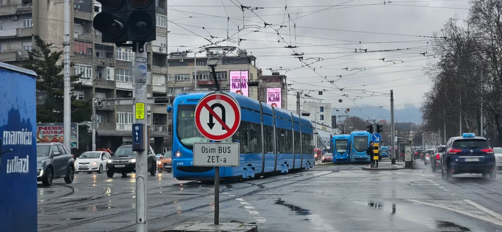 Blue ZET trams and cars move through a wet Zagreb street beneath overhead wires.